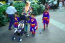 superkids!: twins and a blurred brother wearing superman costume, muslim mother in traditional headdress, baby in pram looking up, panned shot on orchard road